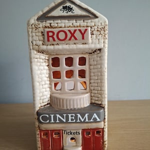 Op de afbeelding: Een keramisch Roxy Cinema-gebouw met een bakstenen gevel. Het gebouw heeft een rood "ROXY"-bord, een "CINEMA"-bord en een loket. Het gebouw is wit, met rode en grijze accenten. Er zit een licht in het gebouw.