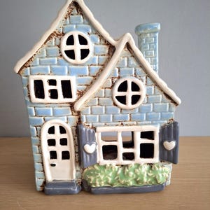 May include: A ceramic house-shaped candle holder with a blue brick pattern. The house has white trim, a chimney, and shutters. The windows are black, and the door is white. It sits on a wooden surface.