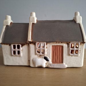 May include: A ceramic cottage with a gray roof, white walls, and a brown door. The cottage has two chimneys and three windows. A white sheep with a black head is on the front. The windows appear to be lit.