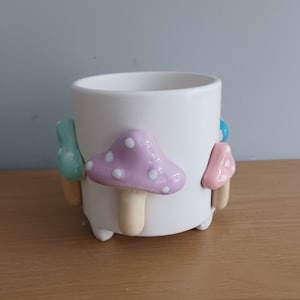 May include: White ceramic planter with mushroom-shaped legs. The planter has a light pink mushroom with white dots on the front. The other legs are light blue, light green, and pink.