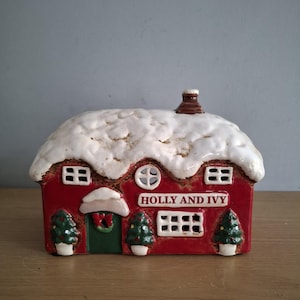 May include: A ceramic house figurine with a red exterior, a white snow-covered roof, and a brown chimney. The front features a green door with a wreath, three small potted Christmas trees, and a sign that reads HOLLY AND IVY.