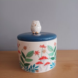 May include: White ceramic jar with a blue lid and an owl finial. The jar has a white background with a forest scene featuring green leaves, red mushrooms, and gold accents.