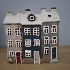 May include: A ceramic tea light holder shaped like a row of three narrow houses. The structure features a white roof, a light grey house with a red door, a dark blue house with a white door, and a teal house with a dark blue door, all with cut-out windows.