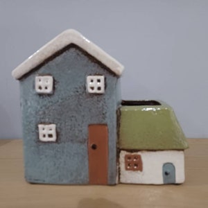 May include: A ceramic house-shaped planter with a blue-gray main structure, a brown door, and white-framed windows. The roof is white, and a smaller section has a green roof and a white and blue door. A charming decorative piece.