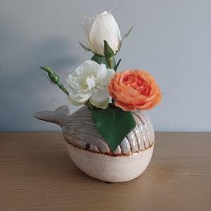 May include: A ceramic whale-shaped vase in cream and gray, holding a small bouquet of flowers. The flowers include a white rose, a white peony, and an orange rose. The vase is on a wooden surface.