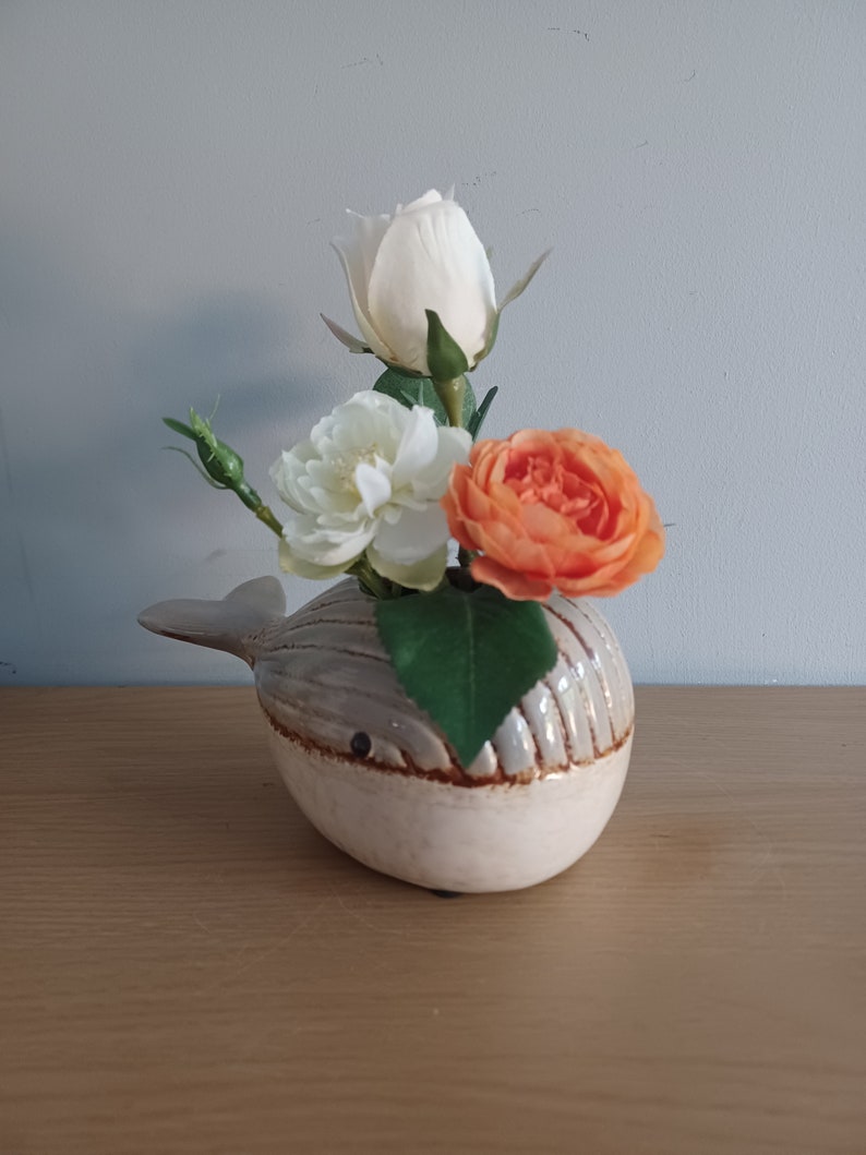 May include: A ceramic whale-shaped vase in cream and gray, holding a small bouquet of flowers. The flowers include a white rose, a white peony, and an orange rose. The vase is on a wooden surface.