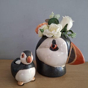 May include: Ceramic puffin-shaped candle holder and vase. The vase is black, white, and orange, with a bouquet of white and peach flowers. The candle holder is a smaller version of the vase, with a candle inside. Both are on a wooden surface.