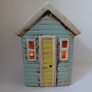 May include: A ceramic beach house figurine with a light blue exterior, yellow door, and windows with orange trim. The house has a white roof and a distressed look.