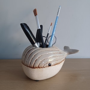 May include: A ceramic whale-shaped desk organizer in cream with brown accents. The whale has a small eye and holds pens, pencils, and paintbrushes. The organizer sits on a wooden surface.