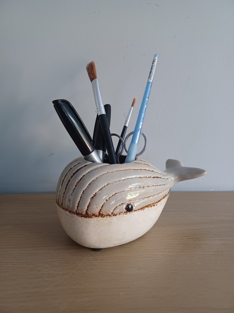 May include: A ceramic whale-shaped desk organizer in cream with brown accents. The whale has a small eye and holds pens, pencils, and paintbrushes. The organizer sits on a wooden surface.