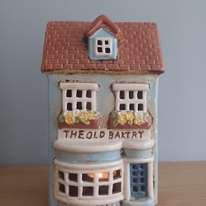 May include: A ceramic house shaped like a bakery with a blue facade, brown roof, and a lit candle inside. The sign reads "The Old Bakery".