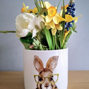 May include: White ceramic vase with a bunny wearing glasses illustration. The vase contains artificial flowers: yellow daffodils, white peonies, and blue hyacinths. A decorative item for the home. The vase is approximately 15 cm tall.