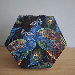 May include: Hexagonal box with a black background, embroidered with two blue peacocks and vibrant tail feathers. The box has a textured surface, possibly for storage or decorative purposes. The design includes peacock feathers.