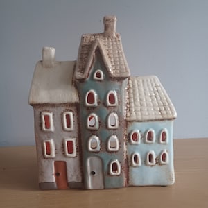 May include: A ceramic house-shaped candle holder featuring three connected buildings. The buildings are painted in grey, teal, and light blue, with cream roofs and chimneys. Each building has multiple windows and a door, with a warm glow visible from the windows.