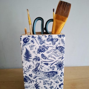 May include: A square wooden pencil holder featuring a white surface with a blue botanical and rabbit pattern. The holder contains several art supplies, including two paintbrushes with wooden handles, a wide brush with gold bristles, and scissors.