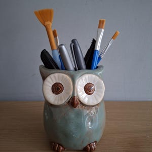 May include: A ceramic owl-shaped pencil holder with a teal glaze. The holder is filled with various art supplies, including paintbrushes and markers.