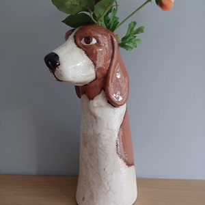 May include: A ceramic vase shaped like a dog's head and neck, with a brown and white colour scheme. The vase holds artificial orange and white flowers with green foliage. The dog's face is detailed, with brown eyes and a black nose.