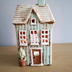 May include: A ceramic house-shaped candle holder with a light blue exterior and a white roof. The house has three windows, a door, and a small chimney. A small potted plant sits to the left of the door. The windows glow with an orange light.