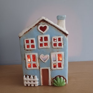 May include: A blue ceramic house-shaped candle holder with a white roof and chimney. The windows glow red, and heart-shaped cutouts are on the front. A small white picket fence and green bush are at the base.
