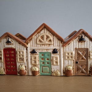 May include: A ceramic planter shaped like three small houses with brown roofs, white walls, and colourful doors and windows. The planter has a rustic farmhouse style.