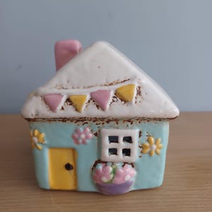 May include: A ceramic house ornament with a light blue body, white roof, and pink chimney. The house has a yellow door, a window, and decorative flowers. The roof is decorated with pink and yellow triangular bunting.