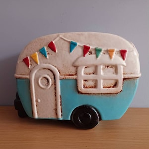 May include: A blue and white ceramic camper figurine with a white door and a window. The camper has a white roof with colorful flags hanging from it.