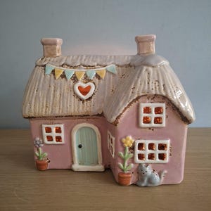 May include: A pink ceramic cottage figurine with a textured thatched roof, a light blue door, and a heart cutout. The house features small windows with orange accents, a colourful bunting banner, potted flower decorations, and a small grey cat figure.