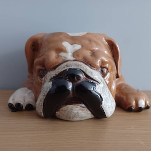 Può includere: Una statuetta di bulldog in ceramica con segni marroni, bianchi e neri. Il bulldog è sdraiato con la testa a terra.