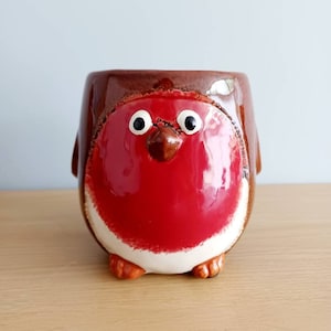 May include: A ceramic robin-shaped mug. The mug has a glossy red breast, white detailing, and brown wings and head. The bird has large, white-rimmed eyes and small orange feet. The mug is displayed on a wooden surface.