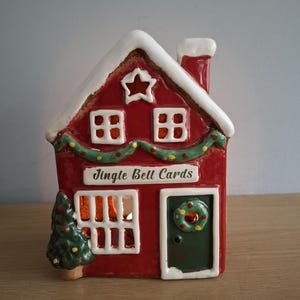 May include: A red ceramic house-shaped tea light holder with a white roof, a chimney, and a green door featuring a wreath. The front displays the text Jingle Bell Cards, a star cutout, two windows, and a small Christmas tree on the left side.