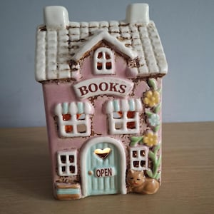 May include: A pink ceramic house-shaped candle holder with the word "BOOKS" above the door. The house has a light blue door with the word "OPEN" and a heart-shaped cutout. A small cat figurine sits near the door.