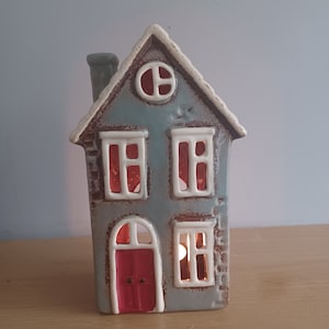 Può includere: Un portacandele in ceramica a forma di casa con una porta rossa e finestre bianche. La casa è dipinta di blu chiaro con accenti marroni.