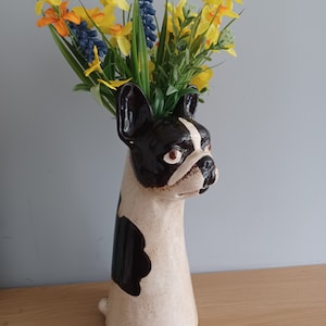 May include: A white and black ceramic vase shaped like a French bulldog with a bouquet of yellow daffodils and blue flowers.