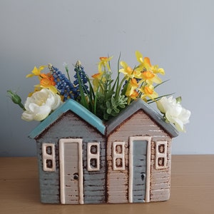 Può includere: Un vaso in ceramica a forma di due case da spiaggia con accenti blu e beige. Il vaso è riempito con un bouquet di narcisi gialli, fiori bianchi e fiori blu.