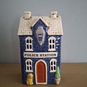 Può includere: Una statuetta in ceramica a forma di stazione di polizia con facciata in mattoni blu, tetto bianco e porta arancione. Presenta una lanterna centrale, quattro finestre, una figura di cane giallo a sinistra e una pianta verde in vaso a destra. La scritta POLICE STATION è stampata in bianco sopra la porta.