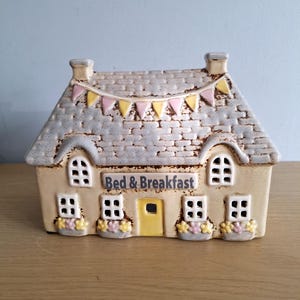 Può includere: Una decorazione a forma di casa in ceramica con un tetto grigio strutturato e uno striscione di bandiere rosa e gialle. La casa ha finestre, fioriere e la scritta "Bed & Breakfast". La casa è beige con una porta gialla.