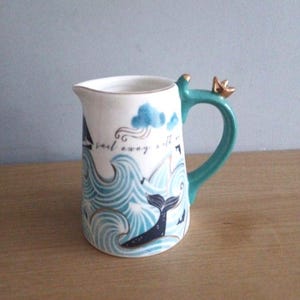 Può includere: Un creamer in ceramica bianca con un manico turchese e una corona dorata in cima. Il creamer presenta un'illustrazione blu e bianca di una coda di balena e onde con il testo "Sail away with me".