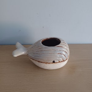 May include: A ceramic whale-shaped planter with a light gray body and white accents. The whale has a brown interior and a small, dark eye. The tail and fins are white. The planter sits on a light brown wooden surface.