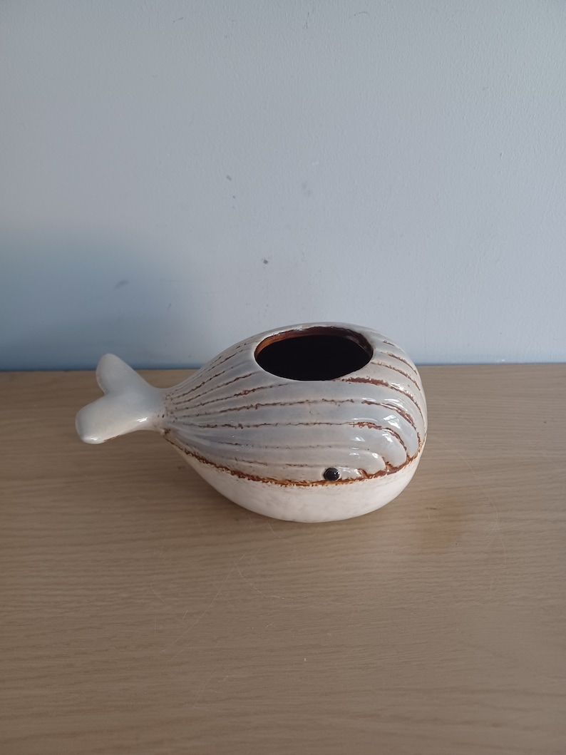 May include: A ceramic whale-shaped planter with a light gray body and white accents. The whale has a brown interior and a small, dark eye. The tail and fins are white. The planter sits on a light brown wooden surface.