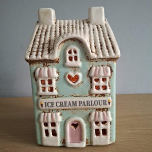 以下が含まれることがあります： アイスクリームパーラーのセラミック製置物。家は水色で、白いトリム、白い屋根、ピンクのドアが付いています。窓はオレンジ色の内装で、ピンクと白のひさしがあります。「ICE CREAM PARLOUR」の文字が前面に表示されています。