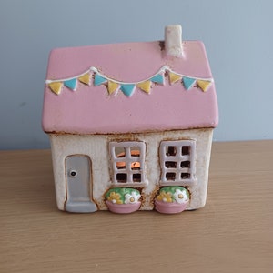 Può includere: Un portacandele a forma di casa in ceramica con tetto rosa e pareti bianche. Il tetto è decorato con una ghirlanda di bandierine blu e gialle. La casa ha due finestre, una porta e vasi di fiori sotto le finestre.