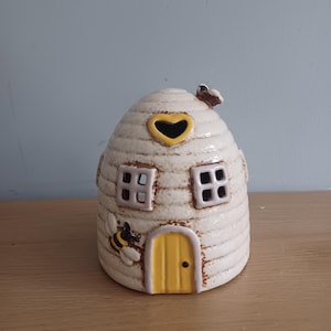 Può includere: Un oggetto decorativo in ceramica a forma di alveare. L'alveare bianco sporco ha una porta gialla, due finestre quadrate e un'apertura a forma di cuore. Un'ape è dipinta sul lato e un'altra in cima.