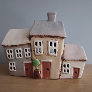 以下が含まれることがあります： 窓とドアが付いた3つの建物がつながったセラミック製の家の置物です。真ん中の建物が最も高く、薄い茶色の屋根をしています。他の2つの建物は白い屋根をしています。真ん中の建物の前に小さな緑色の植物があります。
