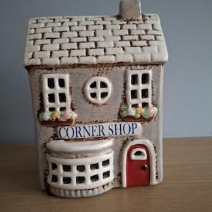 May include: A ceramic Corner Shop ornament. The building features a brick-effect roof, two square windows, and a circular window. The shop front is grey with a red door and a sign reading "CORNER SHOP".