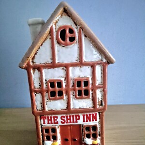May include: A ceramic house ornament with a brown roof, white walls, and brown detailing. The front has windows, a red door, and the text "THE SHIP INN" in red. A chimney is visible on the roof.