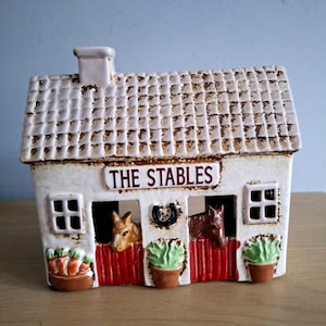 Può includere: Un oggetto decorativo in ceramica a forma di stalla, con tetto e pareti bianche con dettagli marroni. La parte anteriore presenta tre stalle con porte rosse, ognuna con una statuina di cavallo. Il testo "THE STABLES" è visualizzato sopra.