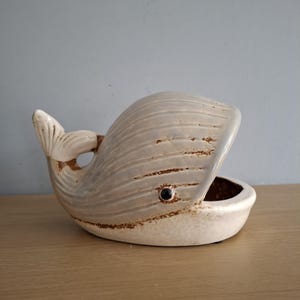 以下が含まれることがあります： 茶色の釉薬と黒い目をつけた白いセラミック製のクジラの形をした皿。クジラの口が開いていて、小さなボウルを形成しています。