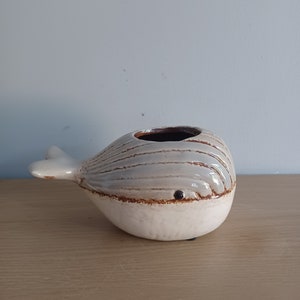 May include: A ceramic whale-shaped vase with a white base and a gray and brown striped body. The whale has a small, dark eye and a fin-shaped tail. The vase has a dark brown interior.