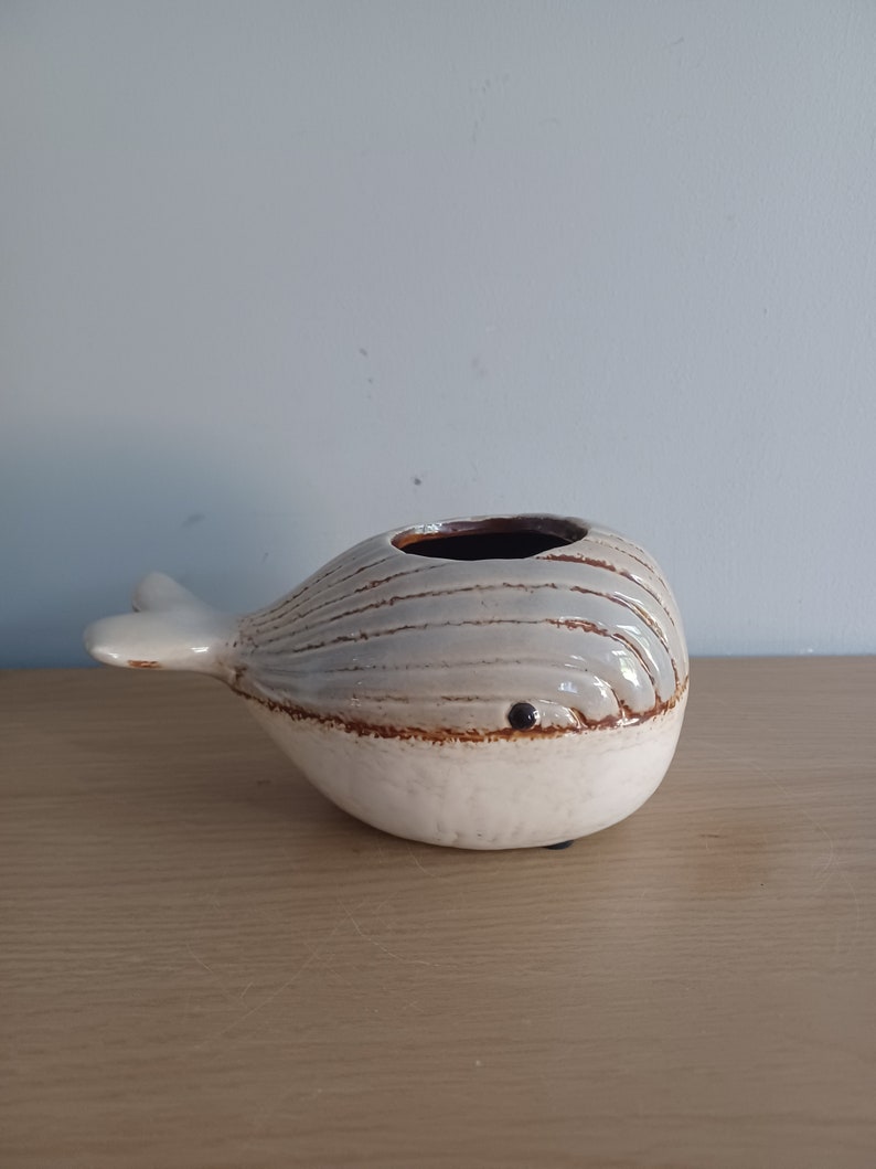 May include: A ceramic whale-shaped vase with a white base and a gray and brown striped body. The whale has a small, dark eye and a fin-shaped tail. The vase has a dark brown interior.