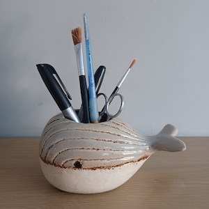 May include: A ceramic whale-shaped desk organizer in off-white with brown accents. The whale holds pens, brushes, and scissors. The organizer is on a wooden surface.